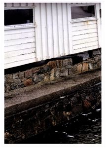 Fotografi som viser nederste del av en hvit bygning med vinduer, støttet opp av en steinmur ved vannkanten.