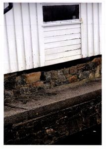 Fotografi av en bygning som viser en hvit fasade med vertikale lister, et vindu, og en steinmur ved grunnmuren.