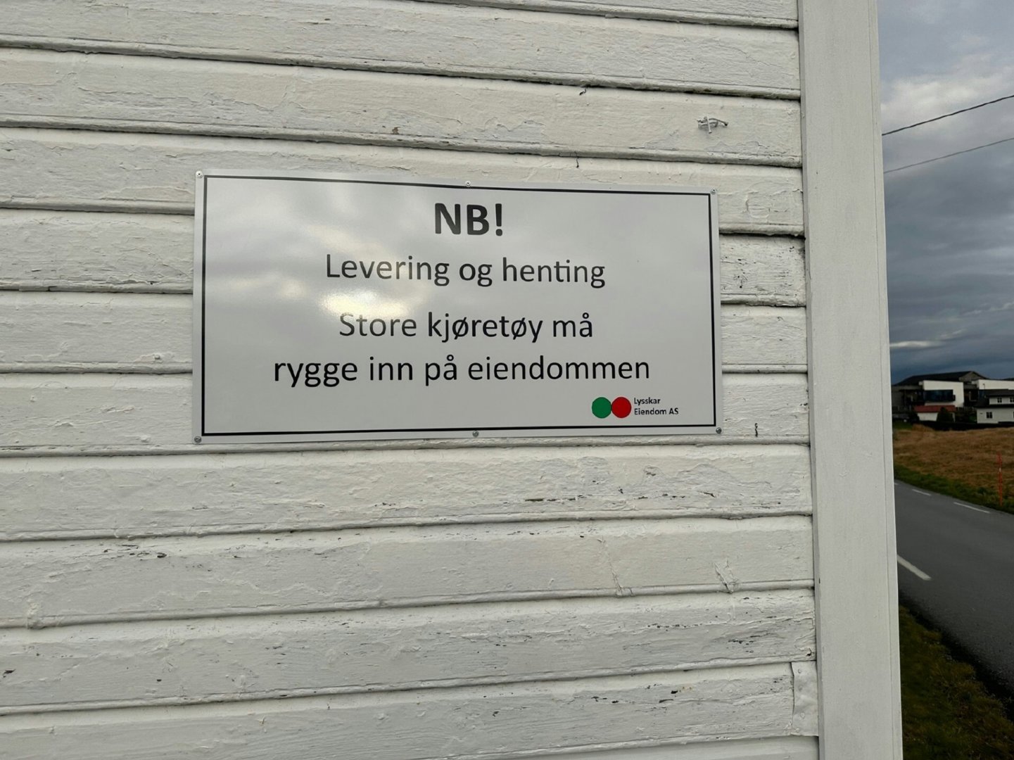 Fotografi av en hvit skilt på en hvitveidet vegggavl. Skiltet inneholder en advarsel om levering og henting av store kjøretøy, samt logoen til 'Eiendom AS'.