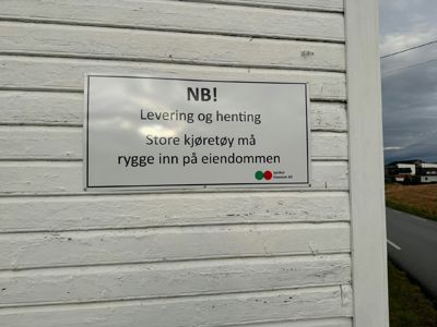 Fotografi av en hvit skilt på en hvitveidet vegggavl. Skiltet inneholder en advarsel om levering og henting av store kjøretøy, samt logoen til 'Eiendom AS'.