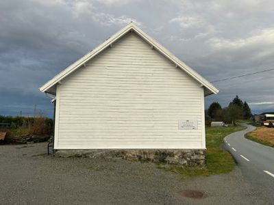 Fotografi av en hvit, trepanelert bygning sett fra siden, med en grusvei og en asfaltert vei i forgrunnen.