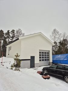 Fotografi av en hvit, moderne bygning (garasje eller uthus) med en stor garasjeport, sett fra siden. Bygningen er omgitt av snø, trær og en bil.