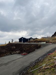 Fotografi av en byggeplass med en ferdig asfaltert vei i forgrunnen og flere mørkeblå hytter på bakken i bakgrunnen.