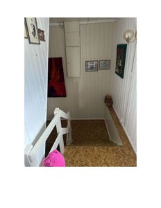 Fotografi av en trapp og et trappeskjulm med hvite panelvegger, gulv av spånplater og diverse innendørsdekorasjoner.