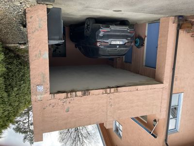 Fotografi av en bil parkert i en garasje eller under et tak, med en rosa fasade i bakgrunnen.