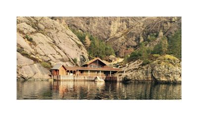 Fotografi av en bygning (trollhytte eller lignende) plassert ved vannet med fjell i bakgrunnen.