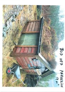 Fotografi av en liten, rustikk hytte eller uthus i en naturlig omgivelser med skog og stein.