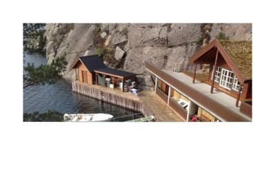 Fotografi av en eksisterende bygning (hus) med en terrasse og en mindre uthus ved vannet, sett fra en høyde.