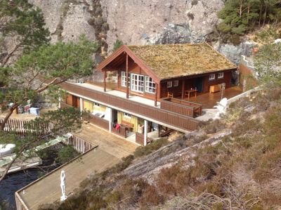 Fotografi av en hytte med grønt tak, plassert på en bratt kystlinje med en brygge og vann i forgrunnen.