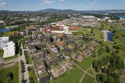 Luftfoto av et boligområde med flere etasjers bygninger, grøntområder, veier og vann i bakgrunnen.