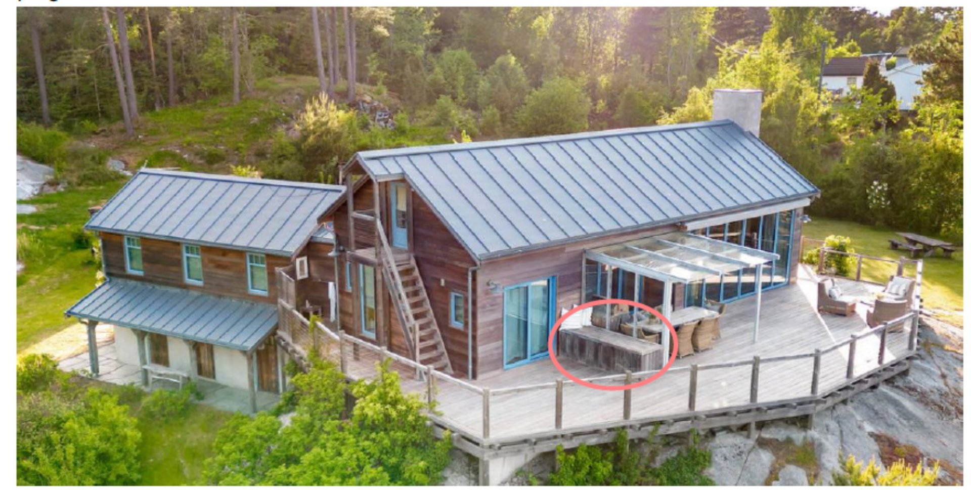 Fotografi av en moderne hytte med trepanel og metalltak, sett fra lufta. Bildet viser bygningens fasade, en stor terrasse med møbler, og omgivelsene med skog og fjell.