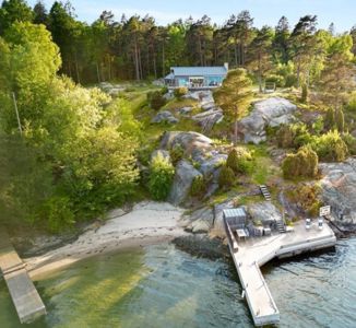 Fotografi av en moderne hytte på en kysttomte med strand, brygge og skog.