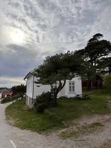 Fotografi av en hvit, trehusbygning med etasjer og vinduer, sett fra siden. Bygningen står på en grønn helling med trær og en brygge eller vei i forgrunnen.