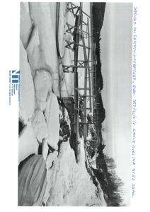 Et svart-hvitt fotografi av en bygning eller konstruksjon i snø. Bildet viser en vinkel fra siden eller bakken.