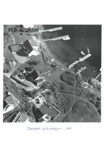 Flyfoto av et kystnært område med boliger, brygger og båter. Bildet viser den fysiske plasseringen av bygningene i forhold til hverandre og til omgivelsene.