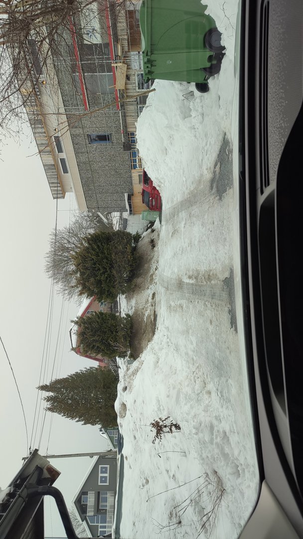 Fotografi tatt fra bil som viser en snødekt vei, nabolagshus og en bygning under oppføring i bakgrunnen.