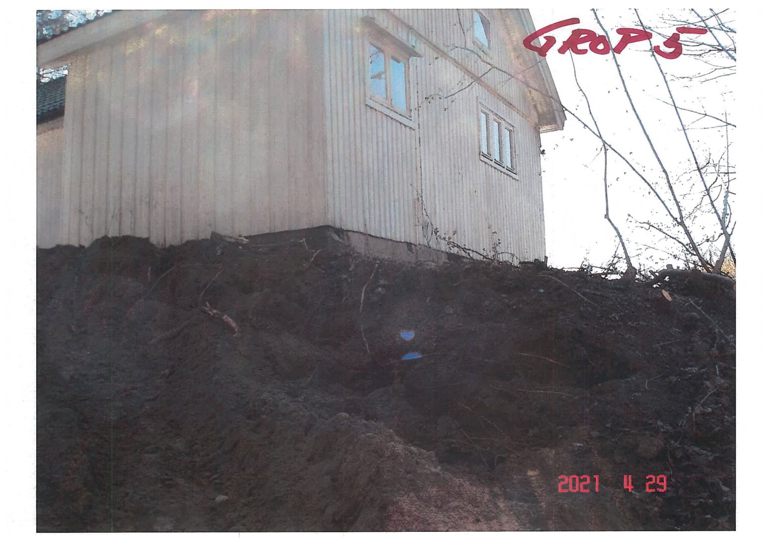 Fotografi av en trehusfasade sett fra bakkenivå, med en steil, jordet bakke i forgrunnen.