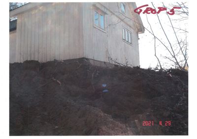 Fotografi av en trehusfasade sett fra bakkenivå, med en steil, jordet bakke i forgrunnen.