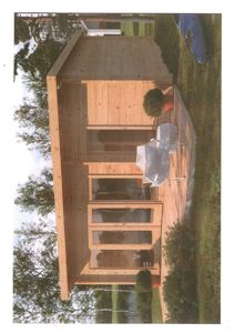 Fotografi av en liten, moderne hytte eller uthus med trepaneling, sett fra siden. Bildet viser bygningens fasade, en liten terrasse med møbler og omgivelsene med trær og gress.