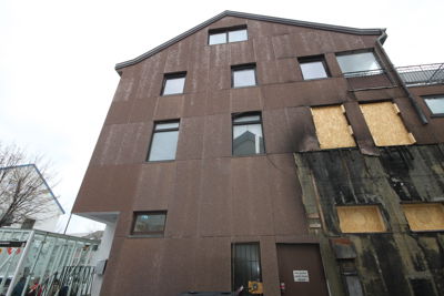 Fotografi av en brun fasade på et fleretasjers bygg, sett fra nede og oppover. Bygget har flere vinduer, hvor noen er dekket med treplater (plywood), noe som antyder at det er under renovering eller reparasjon.