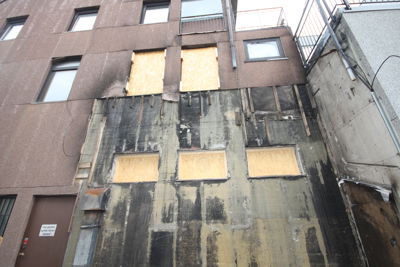 Fotografi av en fasade på et fleretasjes bygg, der vinduene er tildekket med treplater og fasaden viser tegn på skader eller vedlikehold.