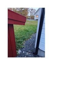 Fotografi av en bygningsside med en svart nedløpsrør (nedløp) som renner ned mot bakken. Bildet viser også en del av en rød bygning i forgrunnen og en grønn hage.
