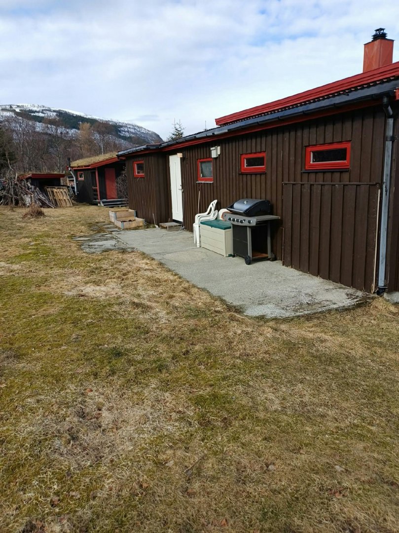 Fotografi av en eksisterende bygning med brun fasade, rød taklist og en grusvei foran. Bildet viser også omgivelser med grøft, en grill og fjell i bakgrunnen.