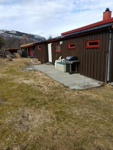 Fotografi av en eksisterende bygning med brun fasade, rød taklist og en grusvei foran. Bildet viser også omgivelser med grøft, en grill og fjell i bakgrunnen.