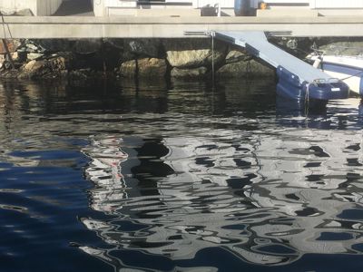 Fotografi av en kai eller brygge som strekker seg ut over vannet, med en båt ved siden av. Bildet viser også refleksjoner i vannet.