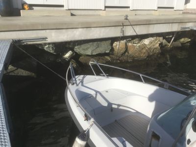 Fotografi av en båt som er festet ved en kai eller brygge. Bildet viser detaljer som vann, steiner under kaien, og båten selv.