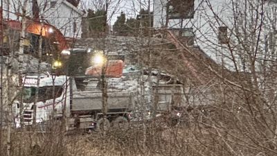 Fotografi av en byggeplass med en lastebil og graver i bakgrunnen, sett gjennom trær.