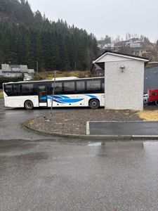 Fotografi av en bussholdeplass eller parkeringsområde med en buss, en liten hvit bygning og en bakgrunn med skog og boliger.