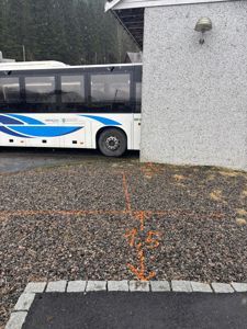 Fotografi av en bussholdeplass eller parkeringsplass med en buss parkert ved siden av en bygning. Det er tegnet oppmåling med oransje maling på grusflaten.