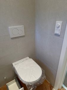 Fotografi av en toalettromsinnredning med toalett, veggmontert flush-knapp og betjeningspanel.