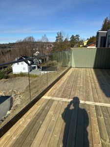 Fotografi av en nybygd terrasse med glassrailing og tredekk, sett fra innendørs eller fra en høyere posisjon. Bildet viser også nabolaget og en annen bolig i bakgrunnen.