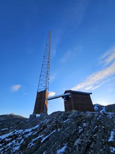 Fotografi av en bygning (tårn og hytte) på en fjelltopp med snø og blå himmel.