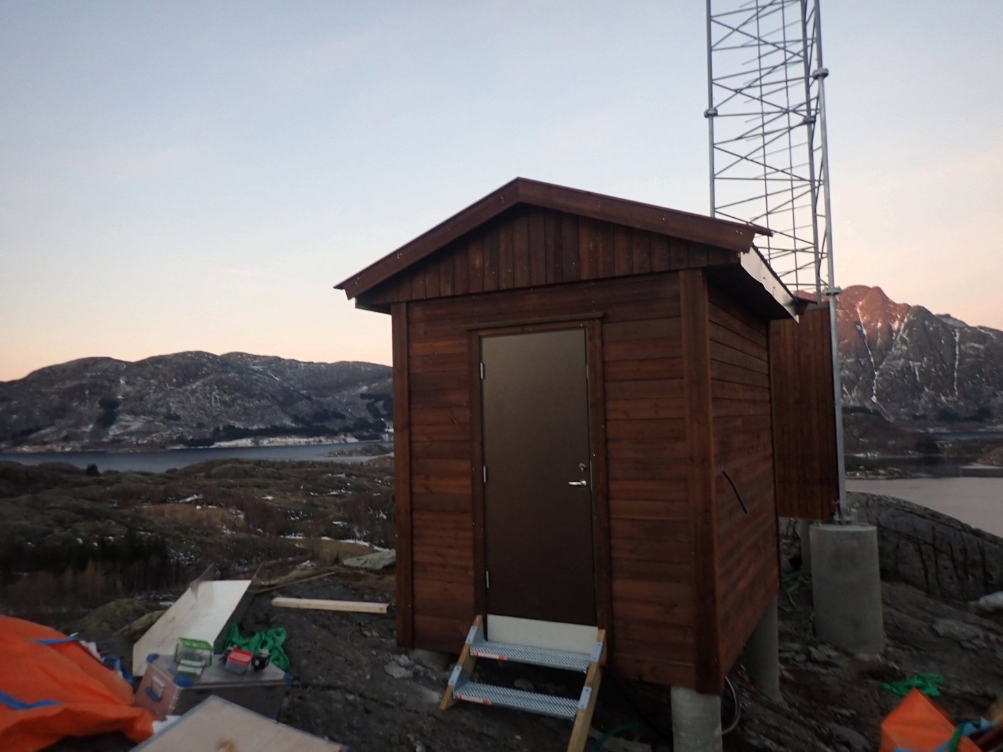 Fotografi av en ferdig eller nesten ferdig bygget liten hytte i tre, sett fra siden med bakgrunn av fjell og vann.