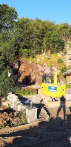 Fotografi av en byggeplass med en gullgraver (CAT) som jobber med å grave ut en steil bakke eller fjellside. Det er også synlig en del av en gul bygning til høyre.