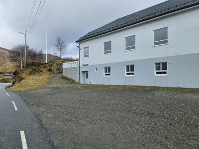 Fotografi av en toetasjes bygning sett fra siden, med en grusplass foran og en vei til venstre.