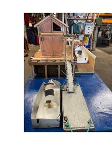 Fotografi av en fysisk modell av en bygning (hus) og tilhørende utstyr, sannsynligvis en modellbåt eller en teknisk prototyp, plassert på en blå arbeidsbenk.