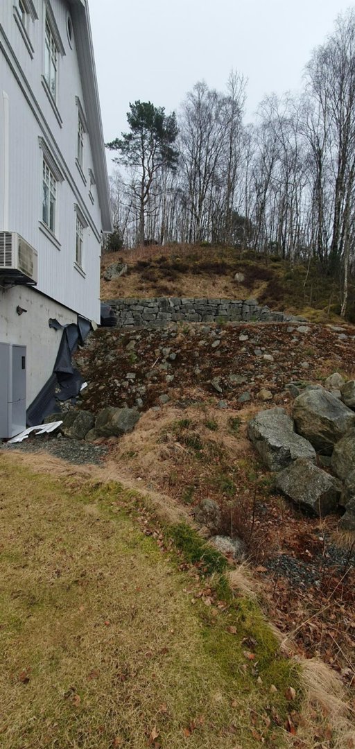 Fotografi av en bygning sett fra siden, med synlig fasade, vinduer, en utendørs klimaanlegg, og terreng med steiner og vegetasjon i bakgrunnen.