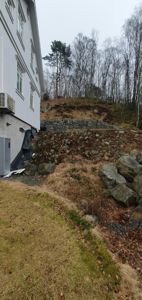 Fotografi av en bygning sett fra siden, med synlig fasade, vinduer, en utendørs klimaanlegg, og terreng med steiner og vegetasjon i bakgrunnen.