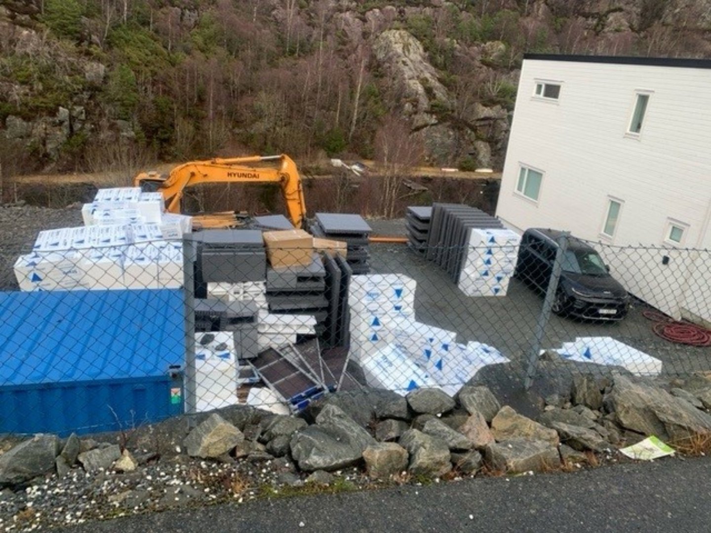 Fotografi av et byggeplassområde med en hvit bygning, en gullfarget Hyundai-graver, en blå container, og en hekk i forgrunnen.