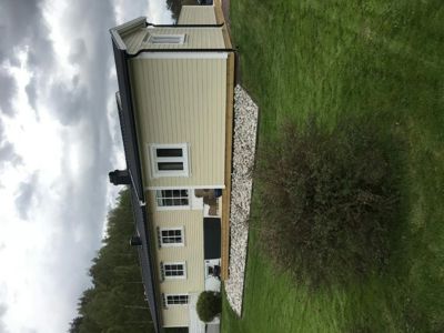 Fotografi av en eksisterende eller ferdigstilt enebolig sett fra siden, med grønt gress og busker i forgrunnen.