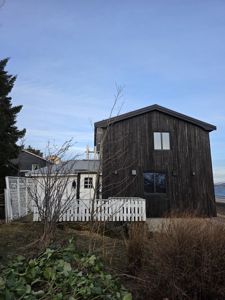 Fotografi av en mørk, trepanelt bygning sett fra siden, med en hvit hekk og vegetasjon i forgrunnen.