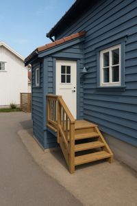 Fotografi av en blå hytte med trapp opp til døren og en hvit hus i bakgrunnen.