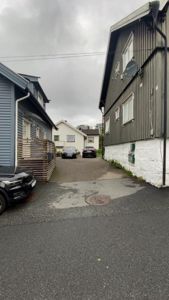 Fotografi av en gate eller innkjørsel mellom to hus, med bilparkering og nabolag i bakgrunnen.