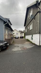 Fotografi av en gate eller innkjørsel mellom to hus, med bilparkering og nabolag i bakgrunnen.