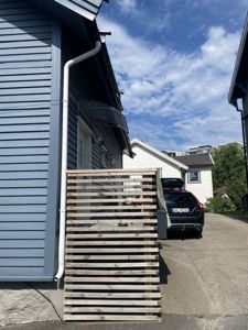 Fotografi av en blå hytte med en hvit nedløpsrør, en trepanelvegg i forgrunnen og en bil parkert i bakgrunnen.