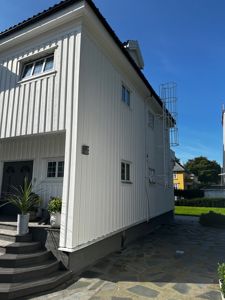 Fotografi av en hvit, toetasje bygning sett fra siden og foran. Bildet viser fasaden, vinduer, trapp opp til inngang, en steinlagt vei og en del av omgivelsene.